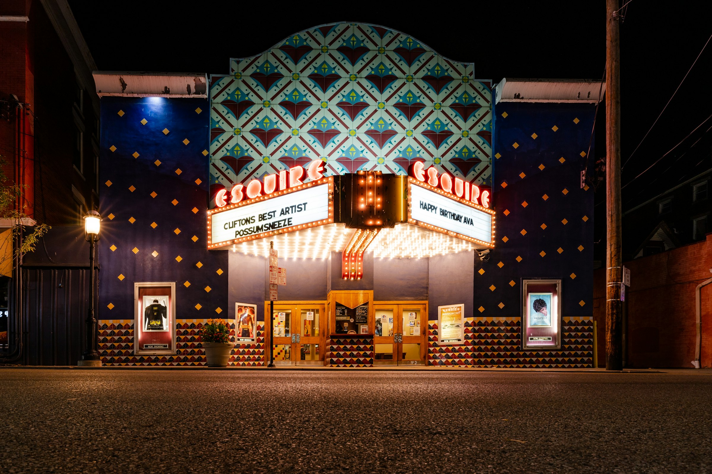 A small-town cinema on a dark street, its marquee bulbs lit and the lobby doors glowing behind them.